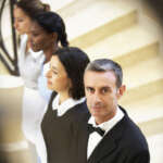 Professional butler standing confidently in front of a line of uniformed household staff on a staircase, representing a well-coordinated domestic team.