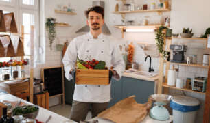 Private chef holding a crate of fresh vegetables in a modern kitchen, symbolizing personalized meal prep for special dietary needs.