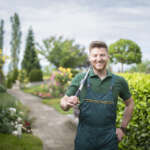 Smiling property manager/estate caretaker standing in a well-maintained garden with hedge clippers, symbolizing seasonal property management and year-round home maintenance.