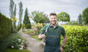 Smiling property manager/estate caretaker standing in a well-maintained garden with hedge clippers, symbolizing seasonal property management and year-round home maintenance.