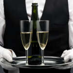 A butler in a black vest and white gloves serves two glasses of champagne on a silver tray, symbolizing refined service and the elevated responsibilities of hospitality staff during the holiday season.