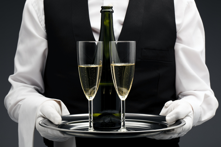 A butler in a black vest and white gloves serves two glasses of champagne on a silver tray, symbolizing refined service and the elevated responsibilities of hospitality staff during the holiday season.