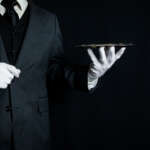 Professional butler in a black tuxedo and white gloves holding a serving tray, representing luxury household service and butler recruitment.