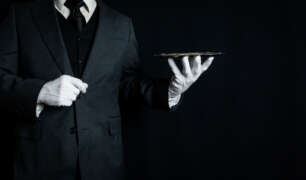 Professional butler in a black tuxedo and white gloves holding a serving tray, representing luxury household service and butler recruitment.