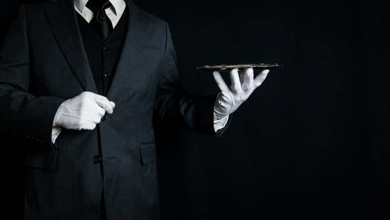 Professional butler in a black tuxedo and white gloves holding a serving tray, representing luxury household service and butler recruitment.