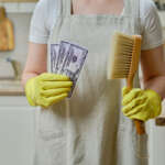 A woman holds money in dollars while cleaning a home kitchen, close-up